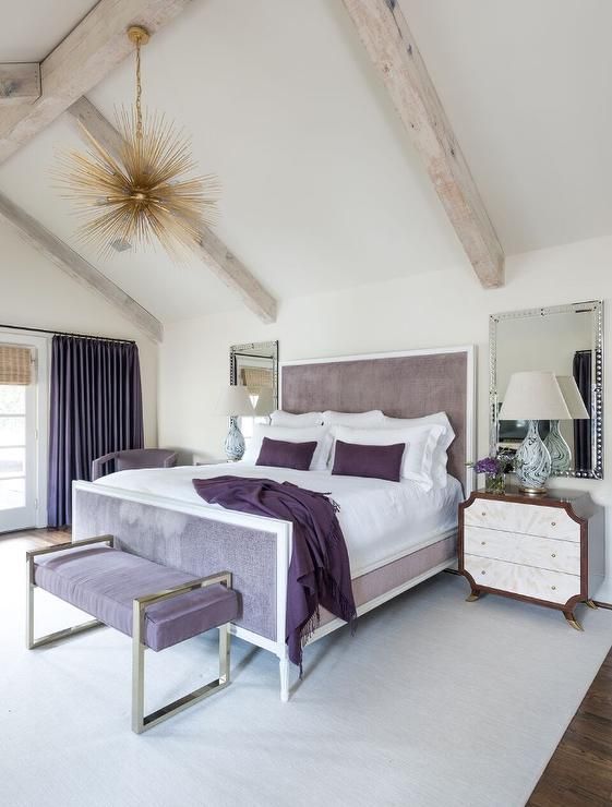 an eclectic bedroom with a lavender bed and an upholstered bench, a sunburst chandelier, wooden beams and purple and white bedding