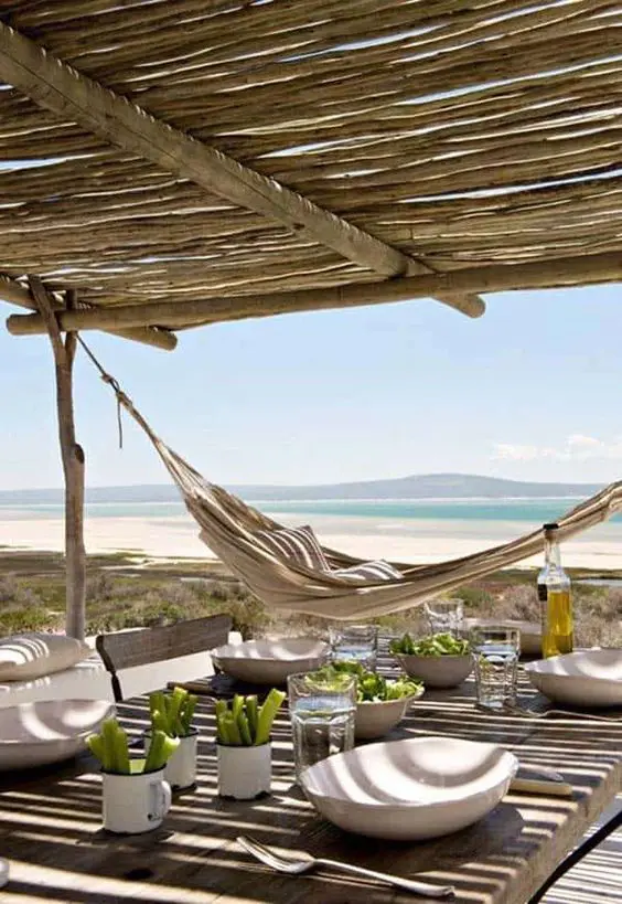 a beach patio with a roof, wooden and wicker furniture, a hammock, some tableware and a gorgeous sea view