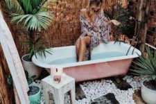 a boho chic outdoor bathroom with pebbles, potted plants, a pink tub and stools and ottomans