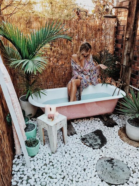 a boho chic outdoor bathroom with pebbles, potted plants, a pink tub and stools and ottomans