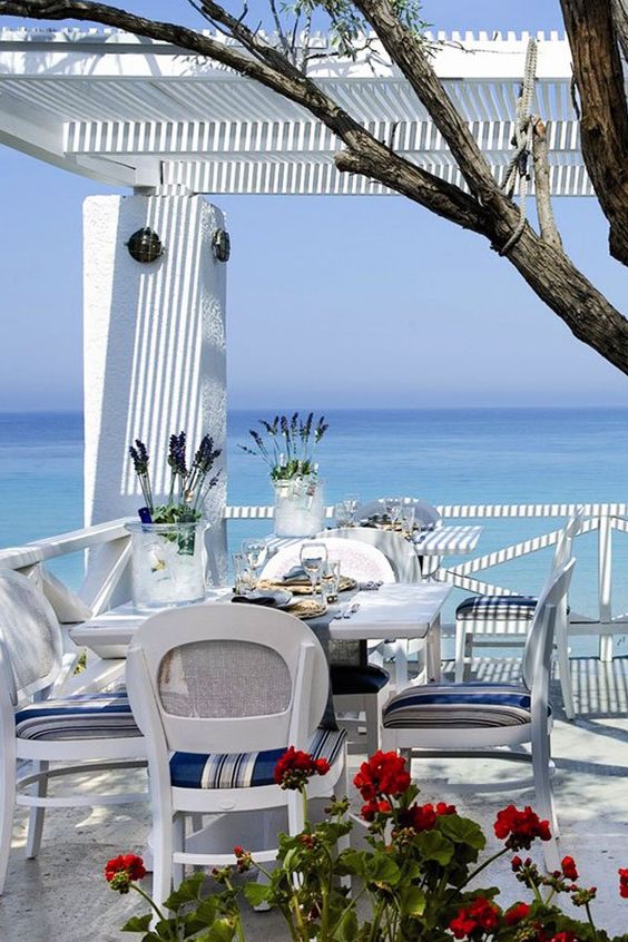 a bright blue and white beach patio with a dining set, bright chairs, some blooms and a tree and a fantastic view