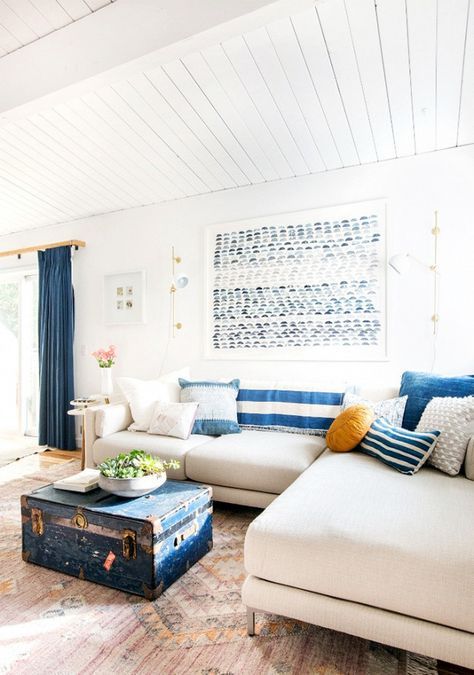 a bright coastal living room with striped pillows, a blue chest, an artwork, blue curtains and a neutral sofa