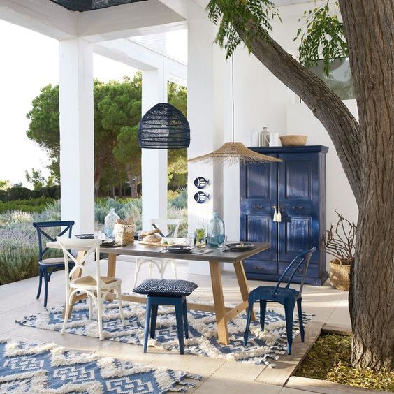 a bright seaside patio in blue and white, with a wooden table and white and navy chairs and stools, pendant lamps and a navy sideboard