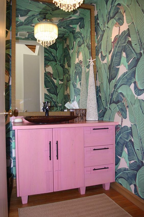 a bright tropical powder room with banan leaf wallpaper, a hot pink vanity, a statement mirror and a vintage chandelier