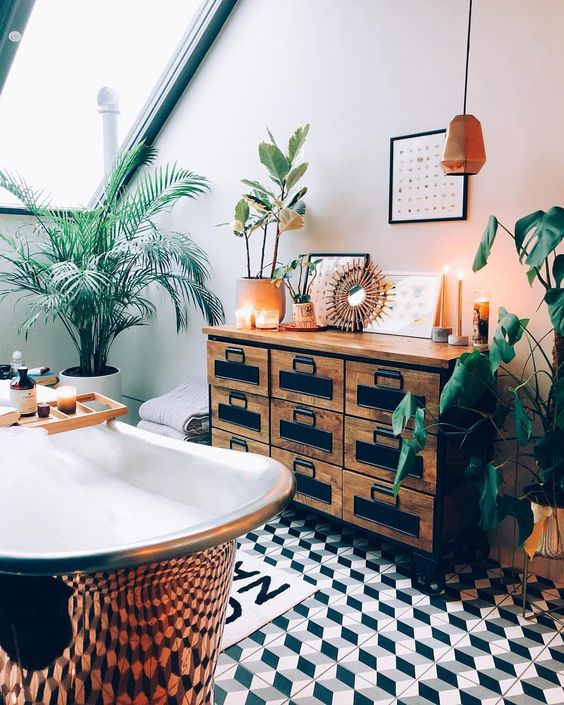 a catchy attic tropical bathroom with a vintage storage unit, a copper clawfoot tub, potted plants and candles plus copper touches