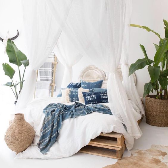 a clean and airy tropical bedroom with a pallet bed with an airy white canopy, wicker pots with green leaves or plants