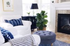 a cozy beach living space with white furniture, a blue ottoman, a printed rug and woven Roman shades
