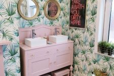 a funny tropical bathroom with tropical wallpaper, a light pink vanity and stools, a neon sign and touches of gold