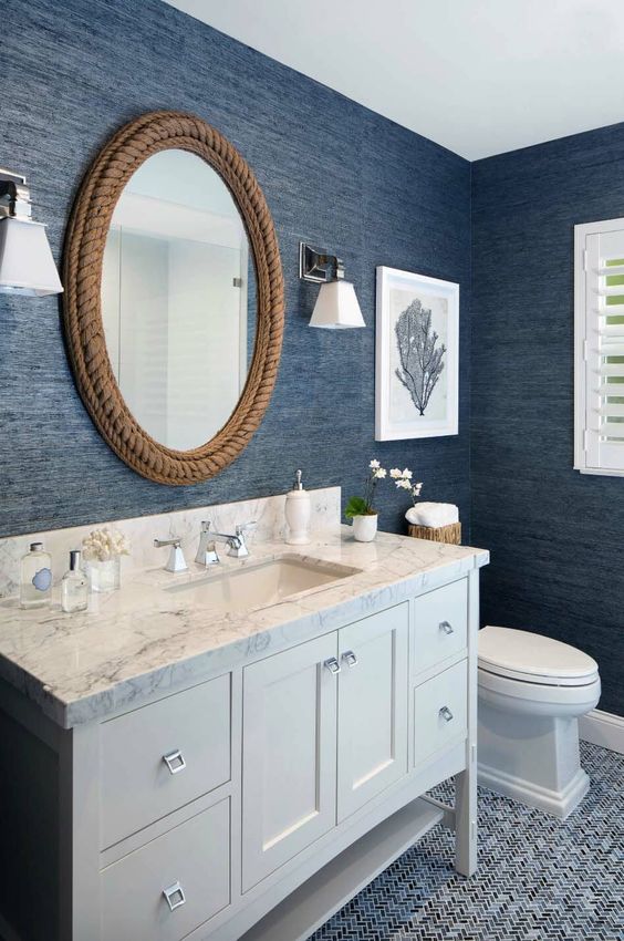 a nautical bathroom with navy textural wallpaper, a rope covered mirror and a white vanity plus artworks