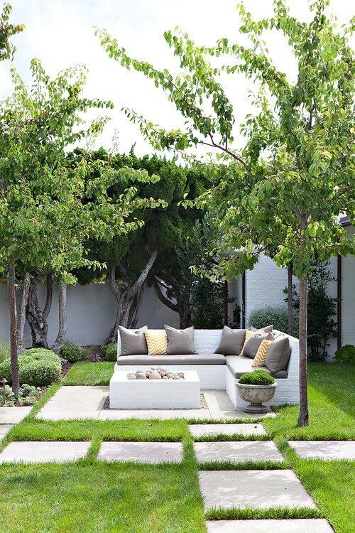 A small and lovely neutral outdoor space with a corner bench with neutral pillows, a box shaped fire bowl with rocks is cool