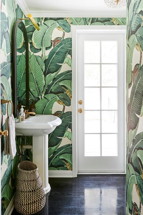 A small tropical powder room with banana leaf wallpaper, a free standing sink, a basket and gold touches