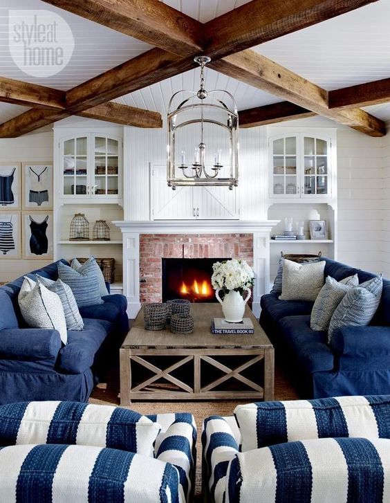 a traditional coastal living room in navy and white, with stripes, a brick clad fireplace, wooden beams and built-in shelves