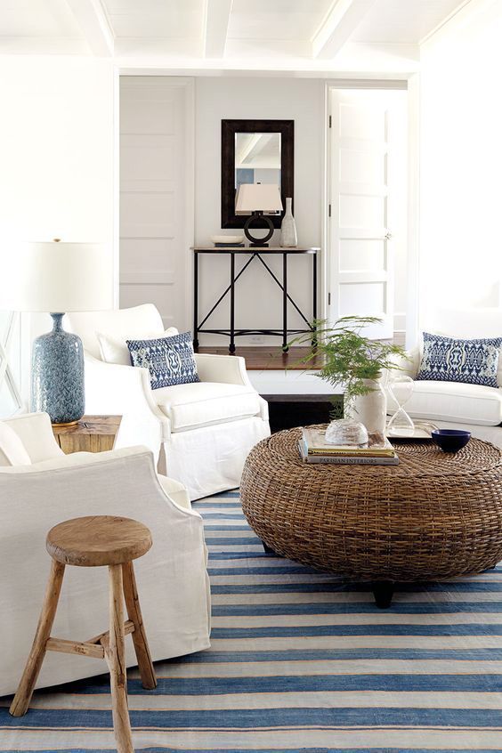 a welcoming coastal living room with a rattan ottoman, a striped rug, white furniture and blue lamps and pillows