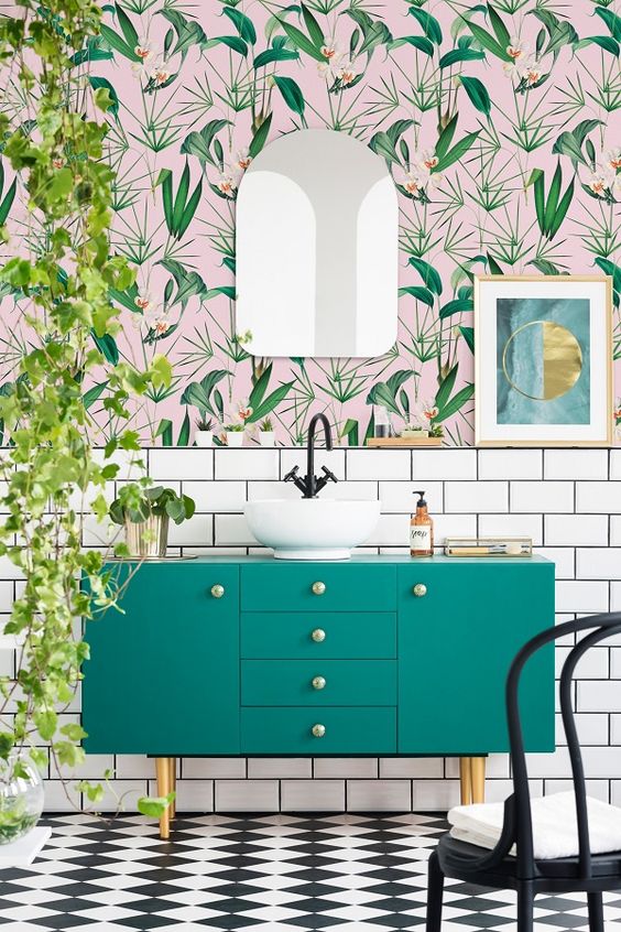 an airy and fun tropical bathroom with catchy wallpaper, a green vanity, cascading greenery and touches of gold