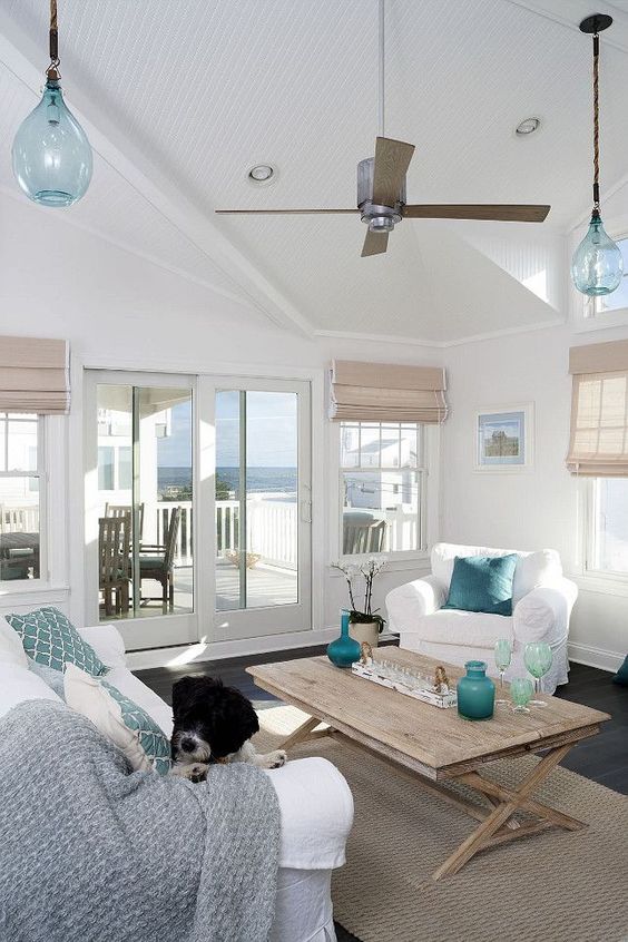 an airy beach living room with woven Roman shades, a wooden table, touches of blue and turquoise, white upholstered furniture