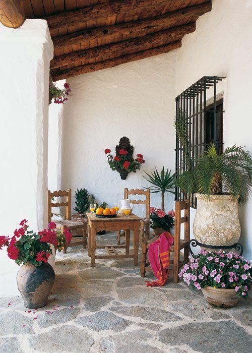 a Mediterranean rustic patio with a stone floor and walls, a wooden ceiling, potted greenery and blooms and wooden furniture