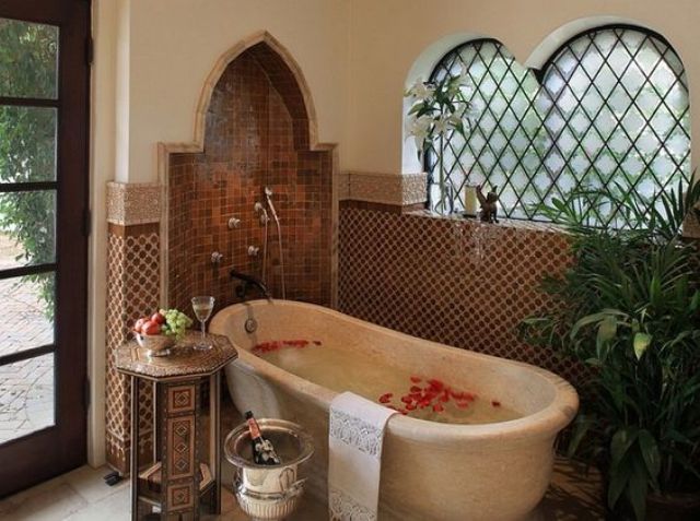 a Moroccan bathroom with catchy tiles, an Eastern-shaped niche, a stone bathtub, a carved side table and potted greenery