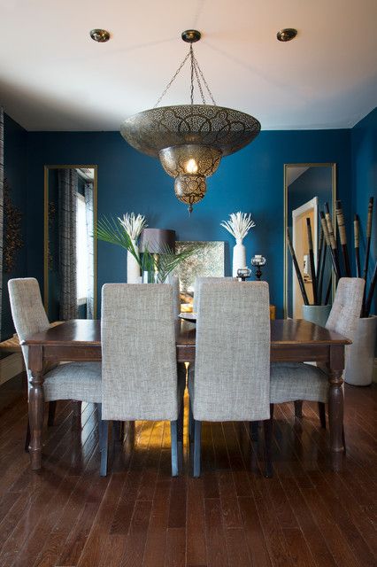 a Moroccan dining room with blue walls, a long table, upholstered chairs, a pendant lamp and a duo of mirrors
