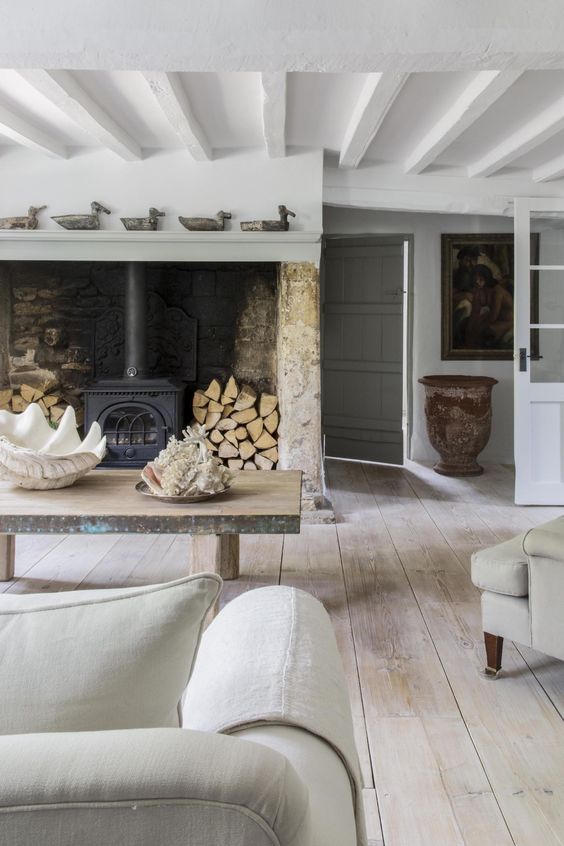 a Provence living room with a hearth with firewood, a rough wooden table and a whitewashed wooden ceiling