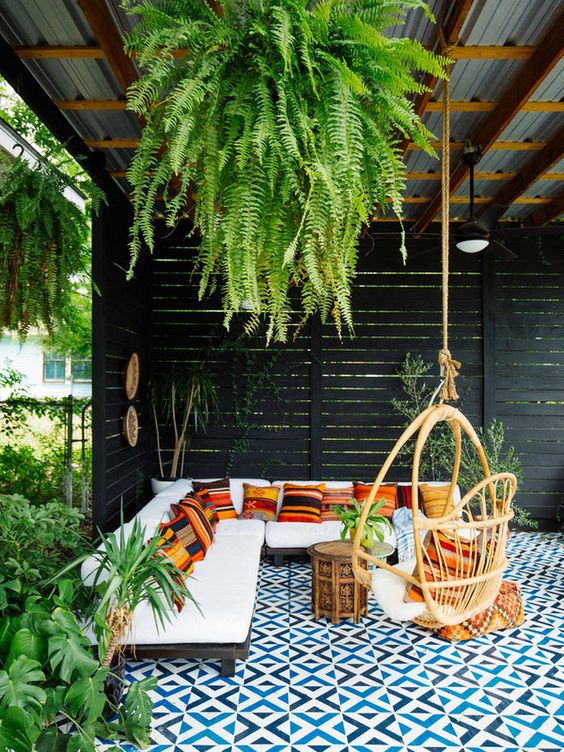 a boho patio with a blue mosaic tile floor, a hanging rattan chair, colorful striped pillows and a bold ottoman