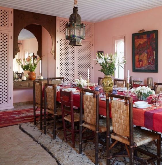 a bold Moroccan dining space with pink walls, a long table with a red tablecloth, woven chairs and a bold pendant lamp