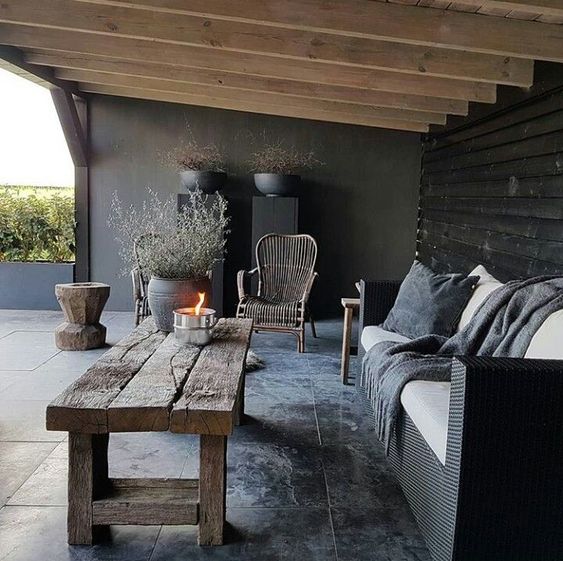 a moody rustic patio with wooden beams on the ceiling, wicker and wooden furniture, potted greenery and minimalist pillows