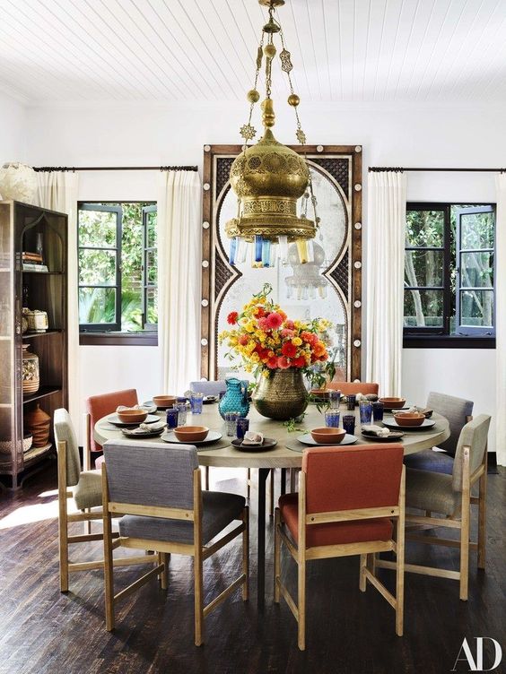 A pretty Moroccan inspired dining room with a round table, muted color chairs, a bold metal pendant lamp and a large mirror in a carved wooden frame