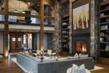 a rustic cabin living room with a wooden ceiling with beams, a stone fireplace and a wall, a large chandelier and neutral furniture