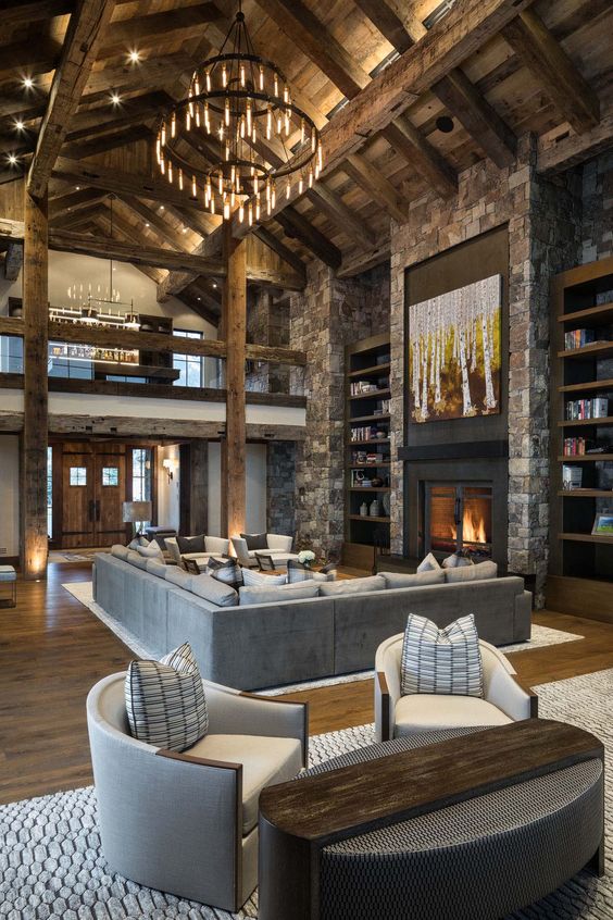 a rustic cabin living room with a wooden ceiling with beams, a stone fireplace and a wall, a large chandelier and neutral furniture