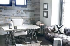 a rustic home office with a salvaged wood wall, a vintage chandelier, a faux fur rug, a trestle desk, a white chair and a leather sofa