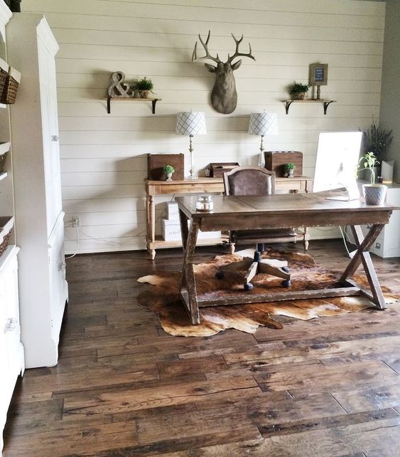a rustic home office with shelves, a wooden console, table lamps, a wooden trestle desk, a leather chair and a deer head