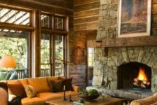 a rustic mountain living room with a stone clad fireplace, a large candle chandelier, cozy furniture and wooden walls