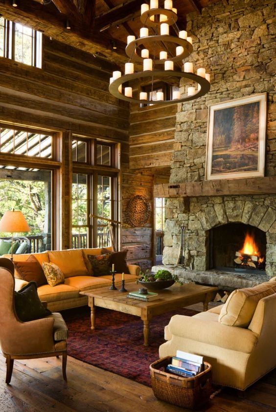 a rustic mountain living room with a stone clad fireplace, a large candle chandelier, cozy furniture and wooden walls