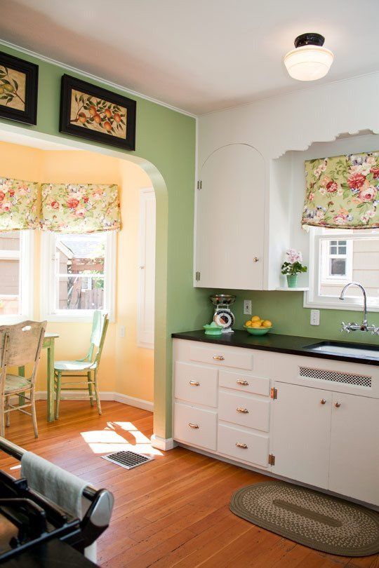 A vintage inspired kitchen with green walls, white cabinets with black countertops, a yellow bay window with green dining furniture