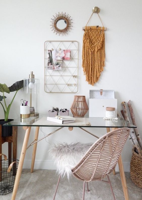 a boho chic home office with a glass desk, a rattan chair, a rattan lantern, a macrame hanging, a sunburst mirror, a memo board and some potted plants