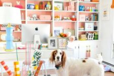 a bright and welcoming home office wiht a large white storage unit with salmon pink backing, a white desk and a plaid rug, colorful accessories and a lamp