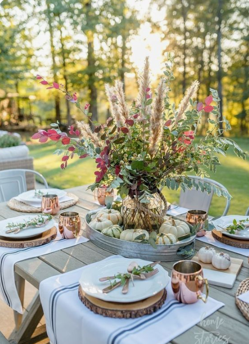 a chic fall tablescape with a greenery, herb and foliage centerpiece, wood slices, copper mugs, striped placemats and natural pumpkins