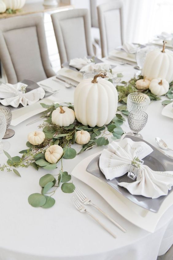a chic white fall tablescape with white linens, white pumpkins, catchy plates, greenery and mini pumpkins