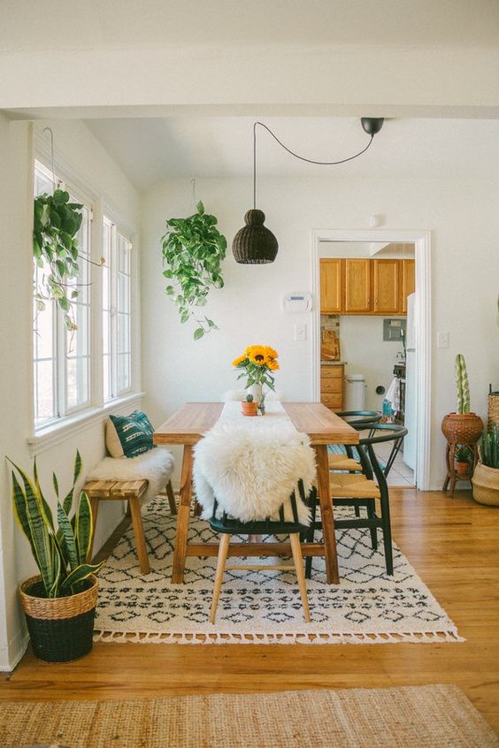 a cozy boho dining nook with simple wooden furniture, a tassel rug, greenery and a wicker lamp hanging over the table