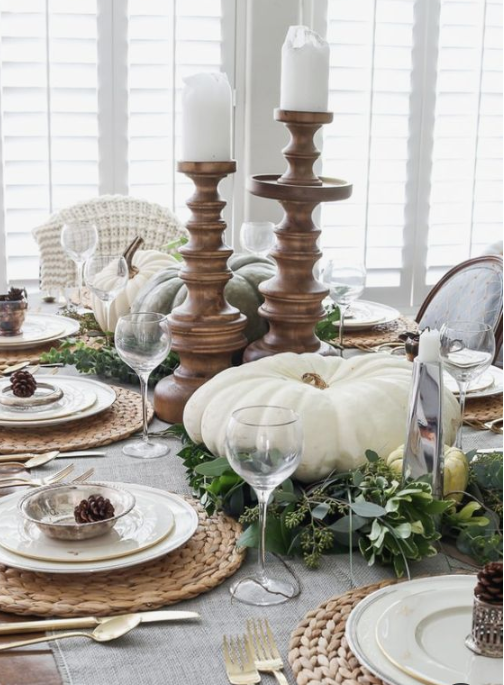 a famrhouse tablescape with heirloom pumpkins, greenery, woven placemats, pinecones and candles in tall wooden candleholders