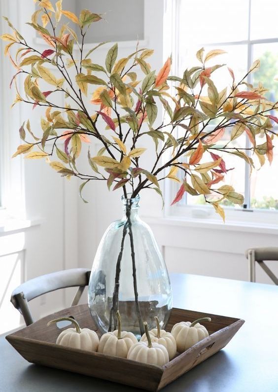 a farmhouse centerpiece of a wooden tray with pumpkins, a glass vase with branches and leaves is a chic and easy idea