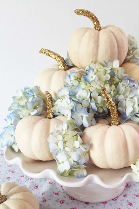 a glam fall centerpiece of a white stand with white pumpkins and gold glitter and blue hydrangeas is chic