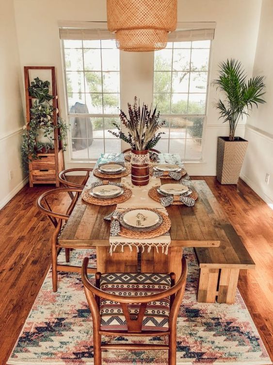 A gorgeous boho dining space with potted greenery, a boho rug, a wicker pendant lamp and warm stained furniture