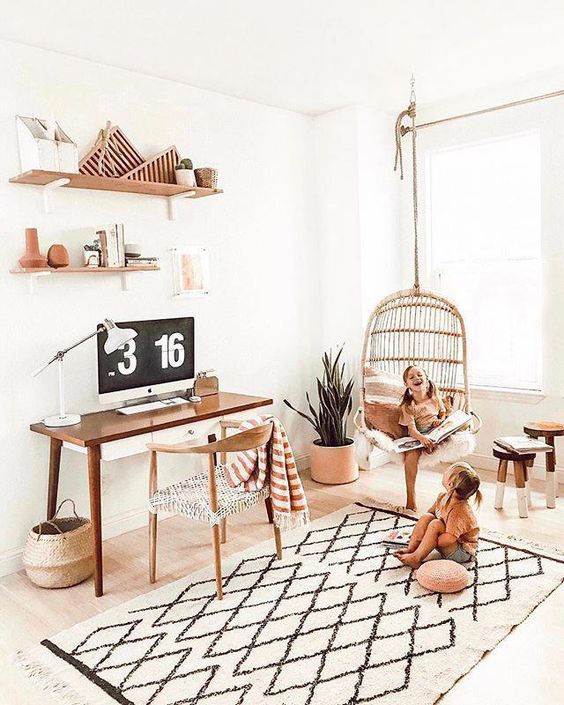 a light-filled home office with a desk and a chair, a printed rug, open shelves, a rattan hanging chair and potted greenery and pillows