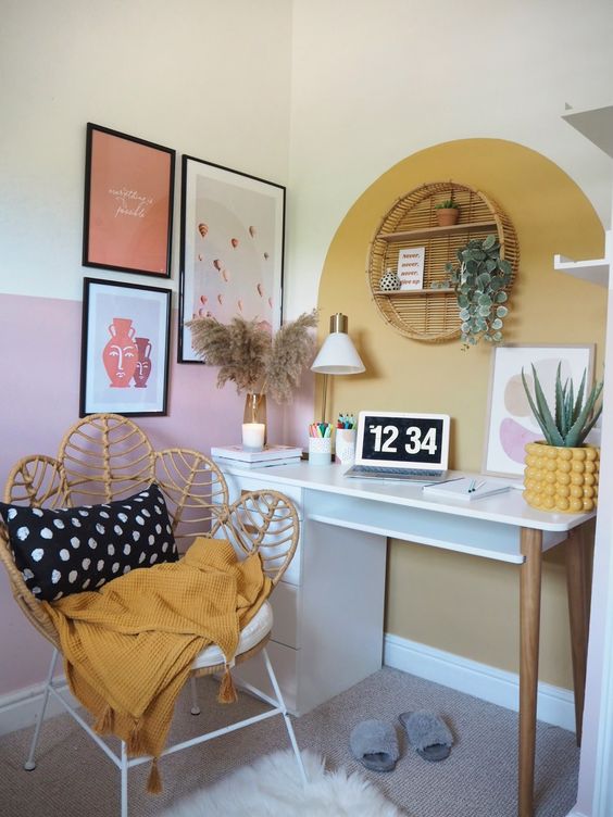 A mid century modern boho home office with a gallery wall, a round rattan shelf, a desk, a rattan peacock chair and pampas grass