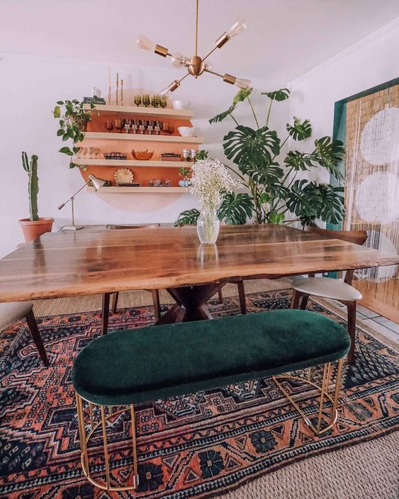 A mid century modern meets boho dining room with a live edge table, a boho rug, a forest green ottoman and potted plants