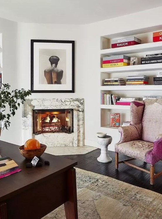 a refined home office with a marble clad fireplace, built-in shelves, a dark stained desk, a pink chair and some art and bright books