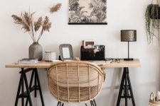 a stylish boho home office nook with a trestle desk, a rattan chair, potted plants and pampas grass, a graphic wall hanging