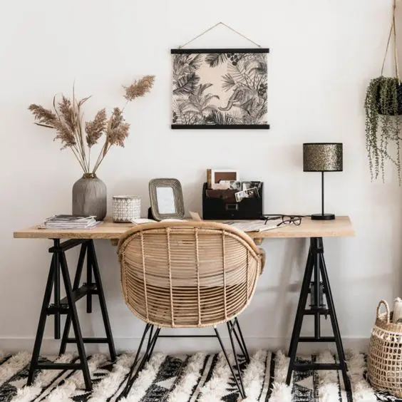 a stylish boho home office nook with a trestle desk, a rattan chair, potted plants and pampas grass, a graphic wall hanging