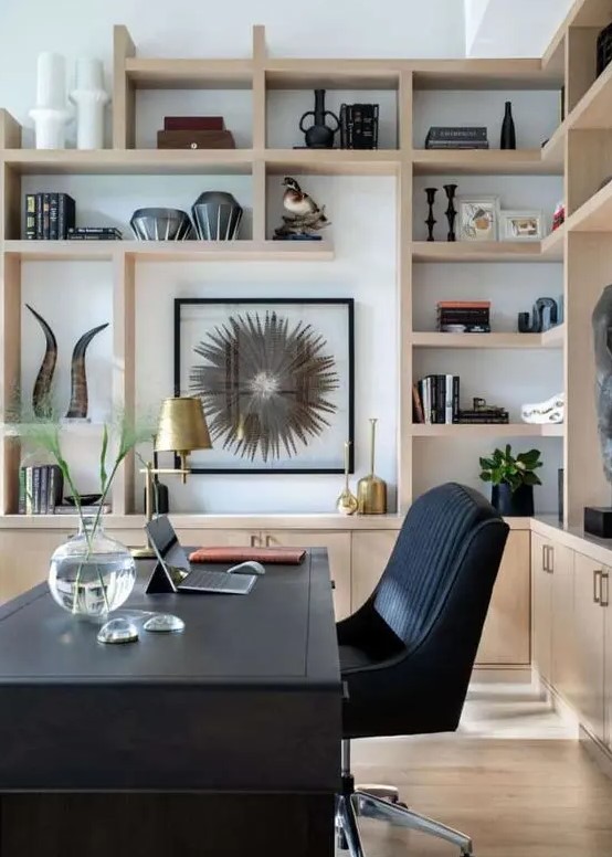 A stylish home office with a black desk and a chair, light stained cabinets and shelves, gilded touches and artwork